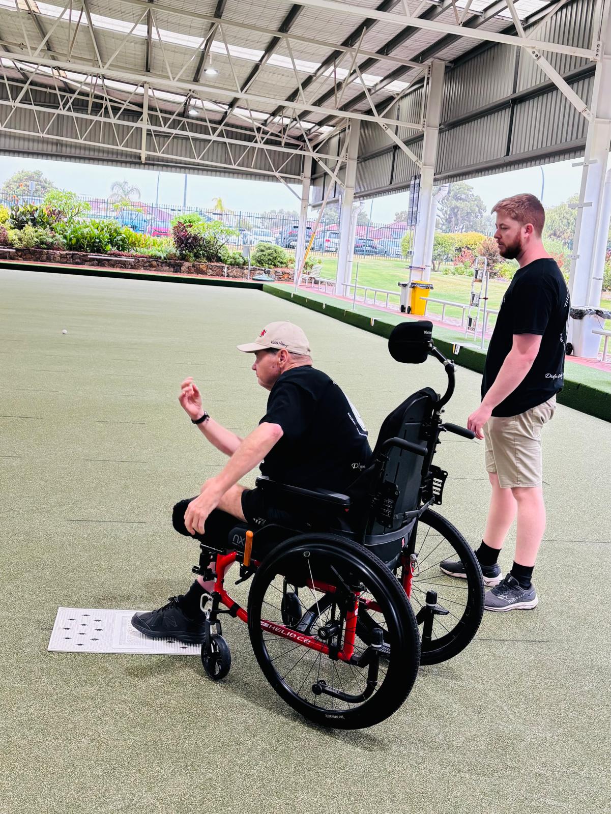 A Happiness Crew member supporting a wheelchair user during a lawn bowls activity